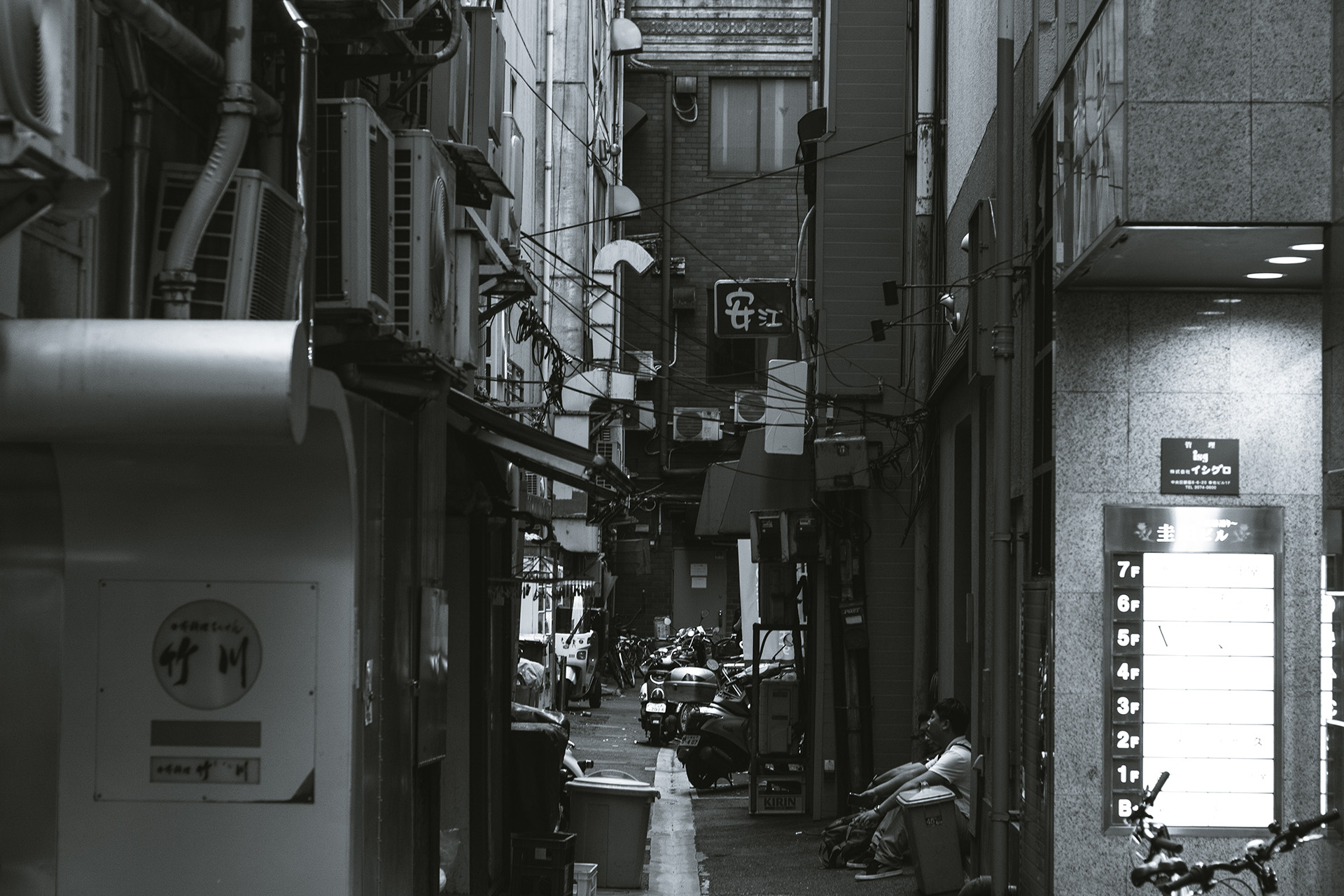 Black and white chaotic and tight alleyway with wires and pipes, two men sitting on the side.
